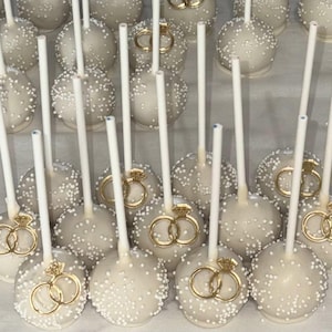 May include: White cake pops with white sprinkles and gold ring decorations on white sticks. The cake pops are arranged in rows, perfect for a wedding or engagement celebration.