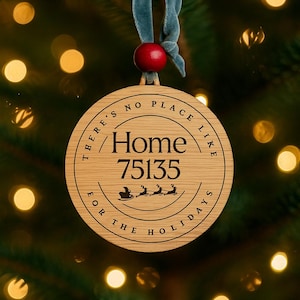 Puede incluir: Adorno navideño de madera con el texto "There's no place like Home for the Holidays" y el número 75135. El adorno es redondo, con una cinta azul y una cuenta roja para colgarlo. El fondo es un árbol de Navidad borroso.