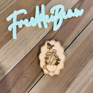 Può includere: Un design in legno tagliato al laser con una tazza da tè piena di fiori e un'ape. Il design è su una superficie di legno. Sopra il design c'è il testo "Freckle Bean" in azzurro.