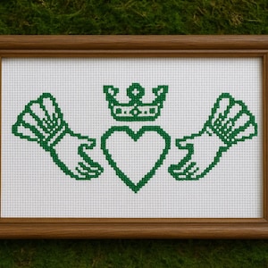 Op de afbeelding: Een ingelijste kruissteek met het Claddagh-symbool, een traditioneel Iers embleem. Het groene ontwerp toont een hart, een kroon en handen op een witte achtergrond. De houten lijst geeft dit stukje Ierse erfgoed een klassieke uitstraling.