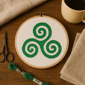 Op de afbeelding: Een afgewerkt kruissteekproject met een groen triskelion-ontwerp, geplaatst in een houten borduurring. Het project is op een witte stof. Groen borduurgaren, een kleine schaar, een kop koffie en een gevouwen doek zijn ook zichtbaar.