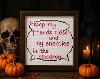 Puede incluir: Un punto de cruz enmarcado con la cita "keep my friends close and my enemies in the cauldron" en hilo rosa y negro. El marco es marrón. La imagen está decorada con calabazas, una calavera, una vela encendida y hojas de otoño.