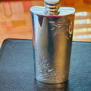May include: A silver-toned metal flask with a polished finish and a screw-top cap. The front surface features an engraved design of a bird in flight near the top and a detailed floral pattern with leaves and stems etched along the bottom corner.