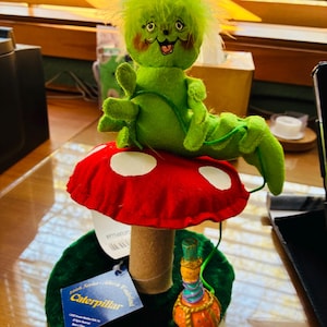May include: A whimsical green caterpillar doll with a smiling face, perched atop a red mushroom with white spots. The mushroom sits on a green base. A small, colorful bottle is next to the mushroom. The doll is part of the 2006 Series Alice in Wonderland collection.