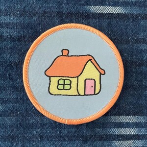 Könnte beinhalten: Ein gestickter Patch in Hellblau und Orange mit einem Cartoon-Haus. Das Haus ist gelb mit einer rosa Tür und grünen Fenstern.