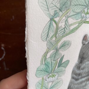 Op de afbeelding: Waterverfillustratie van een grijze tabbykat met een groene klaverkrans. Het kunstwerk bevat gedetailleerde botanische elementen en een zachte, getextureerde papieren rand. De vacht van de kat is weergegeven met delicate schaduwen.