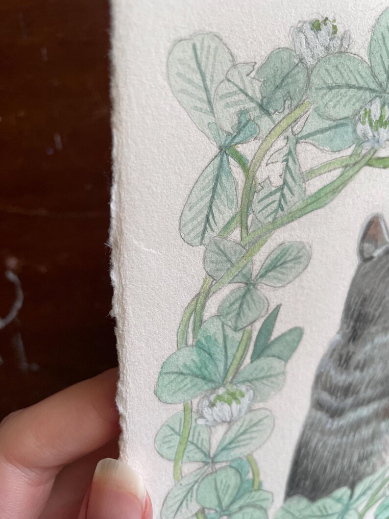 Op de afbeelding: Waterverfillustratie van een grijze tabbykat met een groene klaverkrans. Het kunstwerk bevat gedetailleerde botanische elementen en een zachte, getextureerde papieren rand. De vacht van de kat is weergegeven met delicate schaduwen.