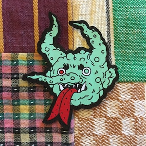 Può includere: Toppa ricamata verde di un mostro dei cartoni animati con una lunga lingua rossa che sporge. Il mostro ha occhi grandi e rotondi e un corpo irregolare e testurizzato.