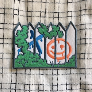 May include: A white picket fence with a blue cross and a dripping orange smiley face in the center. Green bushes are on either side of the fence.