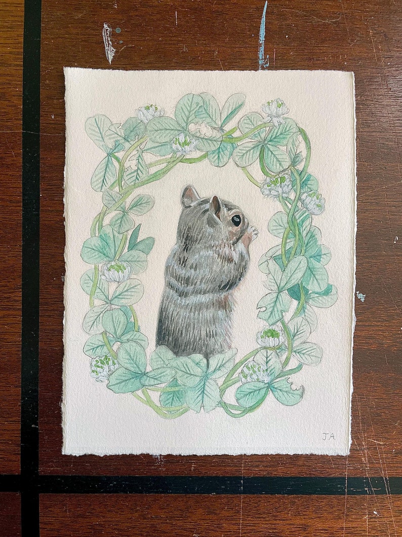 This image is of a painting of a brown, beige, and grey squirrel with its hand toward its mouth surrounded by a wreath of green clover.