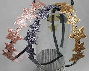 Può includere: Tre fasce per capelli a forma di stella con glitter dorati, rosa e grigi. Le fasce sono su un vaso di vetro trasparente con una base di glitter argentati.