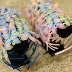 Pu&ograve; includere: Due set di fasce per capelli a forma di farfalla su un espositore nero. Le fasce sono realizzate con nastri blu e rosa con decorazioni a forma di farfalla. Le farfalle sono iridescenti e presentano un arcobaleno di colori.