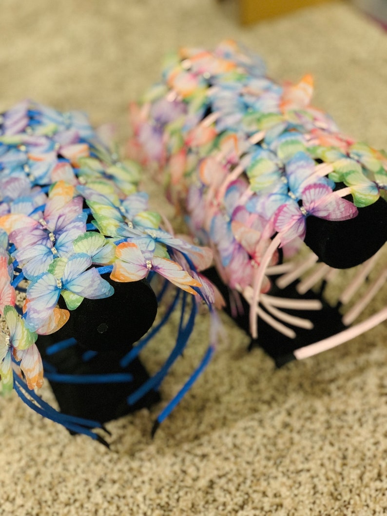Pu&ograve; includere: Due set di fasce per capelli a forma di farfalla su un espositore nero. Le fasce sono realizzate con nastri blu e rosa con decorazioni a forma di farfalla. Le farfalle sono iridescenti e presentano un arcobaleno di colori.