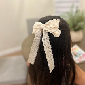 May include: A cream-colored lace bow hair clip. The bow is tied with a long, flowing tail of lace.