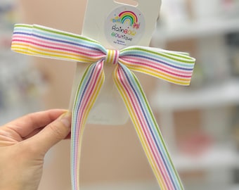 Può includere: Un nastro a righe arcobaleno legato a fiocco con un cartoncino bianco che dice "Rainbow Boutique" con un arcobaleno e un disegno di fiori.