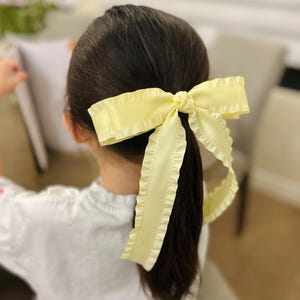 May include: A pale yellow satin ribbon with ruffled edges tied into a large bow, used as a hair tie.