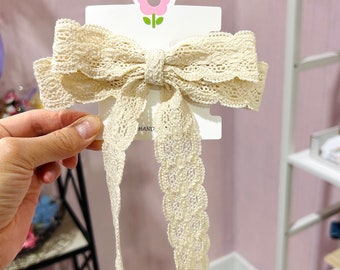 Può includere: Un fiocco bianco all'uncinetto con un lungo nastro abbinato. Il fiocco è attaccato a una carta bianca con un'illustrazione di un fiore rosa e la parola "HAND" stampata su di essa.