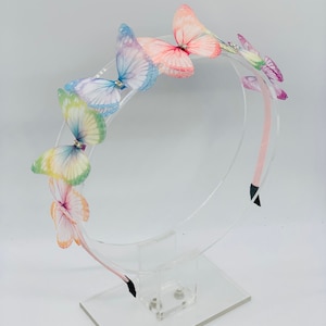 Pu&ograve; includere: Un cerchietto per capelli con una fila di farfalle colorate. Le farfalle sono fatte di tessuto trasparente e hanno ali iridescenti. Il cerchietto &egrave; rosa e ha un elastico nero.