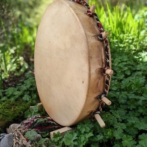 May include: A large, round drum with a light-colored wooden surface and wooden pegs. The drum is decorated with a black, red, and white braided rope. The drum is set against a backdrop of green foliage and natural elements.