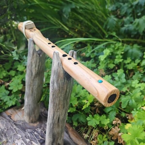 May include: A light-coloured wooden Native American style flute featuring a smooth grain, five black finger holes, a small turquoise inlay near the end, and a leather-bound block secured with a light-coloured cord on the top section.