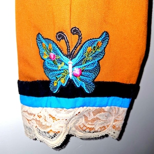 May include: A mustard yellow garment sleeve featuring an embroidered blue butterfly with pink and yellow floral accents. The cuff is detailed with a horizontal black velvet band, a bright blue fabric strip, and a scalloped cream-colored lace trim.