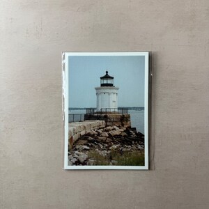 May include: A photograph of a white lighthouse with a black top, situated on a stone pier. The lighthouse is surrounded by rocks and the ocean. The sky is a pale blue. The photograph is in a clear plastic sleeve.