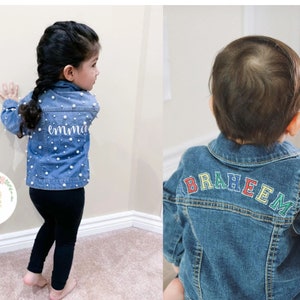 May include: Two children wearing denim jackets with personalized embroidery. The girl's jacket has white pearl embellishments and the name "emmie" embroidered on the back. The boy's jacket has the name "BRAHEEM" embroidered on the back in rainbow colors.