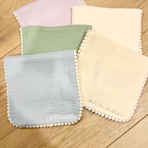 May include: Five soft, square baby bibs in pastel colors: lavender, green, blue, and two shades of cream. Each bib has a white pom-pom trim and embroidered text. The bibs are arranged on a wooden surface.