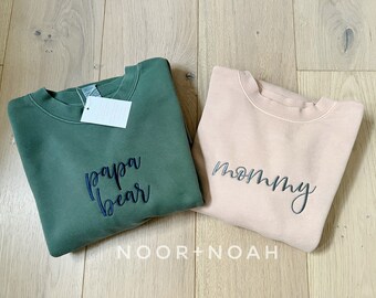 Könnte beinhalten: Zwei Sweatshirts, eines grün und eines hellrosa, mit gesticktem Text. Das grüne Sweatshirt sagt "papa bear" und das rosa Sweatshirt sagt "mommy".