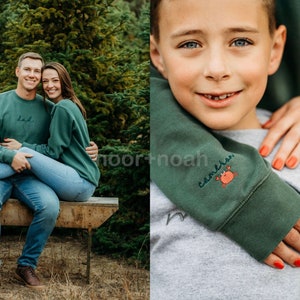 May include: A family of three wearing matching green sweatshirts with embroidered text. The father is wearing a sweatshirt that says "Dad", the mother is wearing a sweatshirt that says "Mom", and the son is wearing a sweatshirt that says "Cameron" with a small crab design. The family is posing in front of a Christmas tree farm.