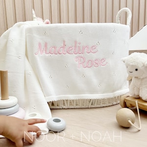 May include: A white woven basket with handles, holding a white blanket embroidered with the name "Madeline Rose" in pink. A plush lamb and a wooden toy are visible. The basket sits on a light wood surface.