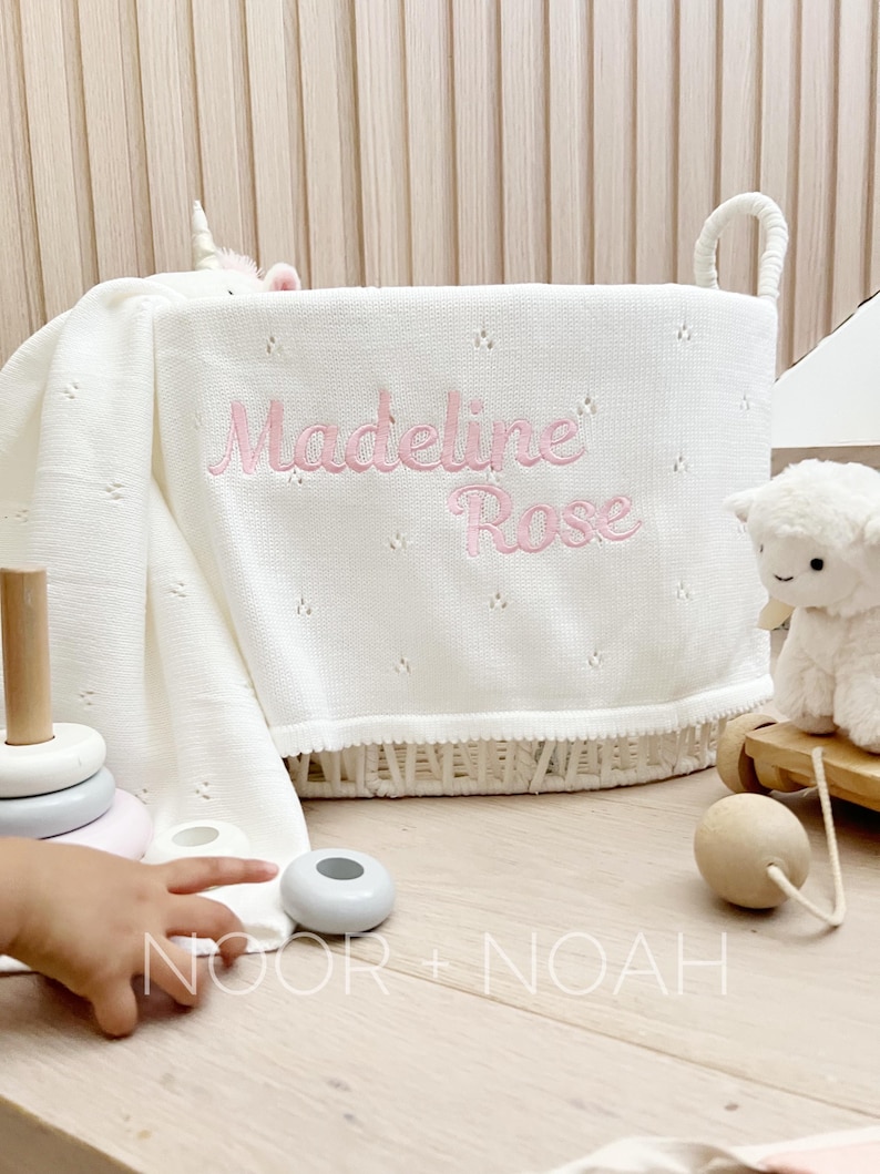 May include: A white woven basket with handles, holding a white blanket embroidered with the name "Madeline Rose" in pink. A plush lamb and a wooden toy are visible. The basket sits on a light wood surface.