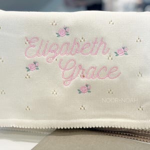 May include: Cream knitted baby blanket featuring the name "Elizabeth Grace" embroidered in pink script, decorated with pink rose motifs. The blanket has a scalloped edge and the brand name "NOOR.NOAH" is at the bottom.