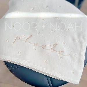 May include: Off-white baby blanket with a scalloped edge and a delicate, dotted pattern. The blanket features the embroidered name "Phoebe" in a cursive font, with the text "NOOR + NOAH" above it. The blanket is made with beige thread.