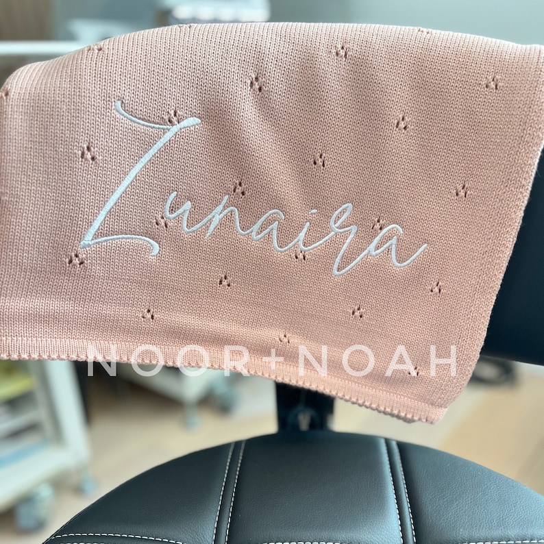 May include: A light pink baby blanket with the name "Zunaira" embroidered in white script. The blanket has a delicate, openwork design and is draped over a black surface. The brand name "NOOR + NOAH" is printed at the bottom.