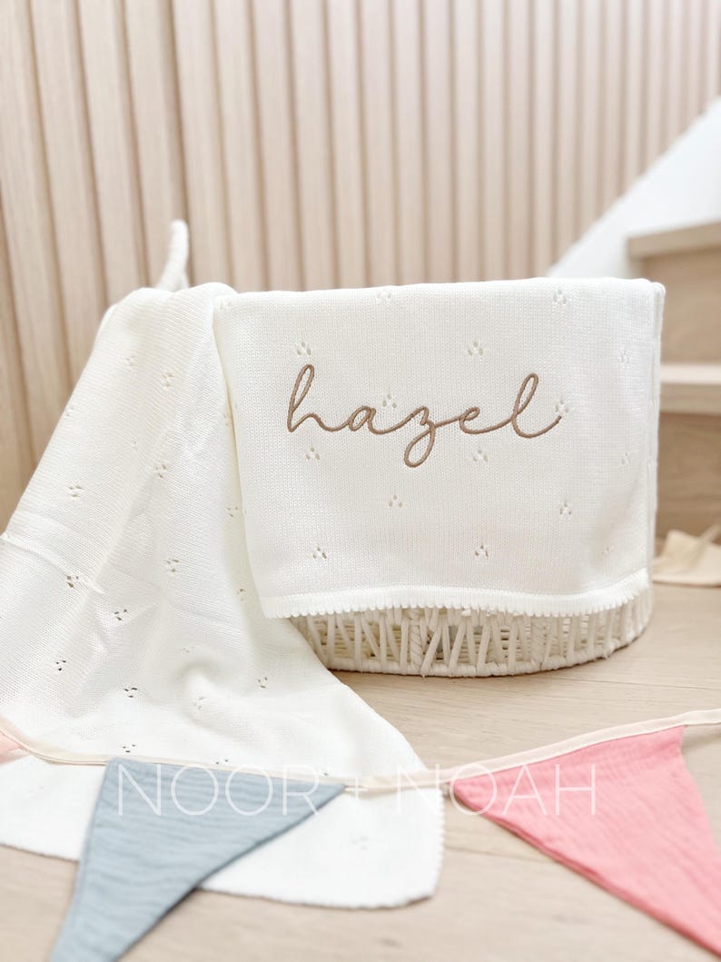 May include: A white knitted baby blanket with the name "Hazel" embroidered in brown cursive script. The blanket is folded over a white wicker basket. A banner with the text "NOOR + NOAH" is visible.