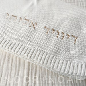 Op de afbeelding: Een wit gebreid babydekentje met gouden Hebreeuwse letters die "דודו אלעד" lezen.