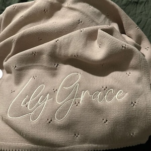 May include: A light pink knitted blanket with the embroidered name "Lily Grace" in white script. The blanket has a scalloped edge and small, evenly spaced decorative holes.