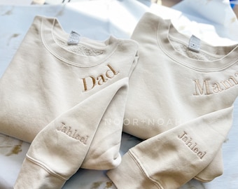 Könnte beinhalten: Zwei beige Sweatshirts mit gesticktem Text. Eines trägt die Aufschrift "Dad" und "Jahleel" auf dem Ärmel, das andere "Mama" und "Jahleel" auf dem Ärmel. Der Markenname "NOOR+NOAH" ist sichtbar.