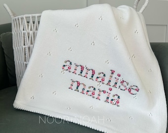 May include: A white knitted baby blanket featuring a delicate pointelle pattern and the name "annalise maria" embroidered in a floral print. The text "NOOR & NOAH" is visible in the lower portion of the blanket fabric.