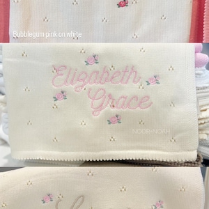 May include: White baby blankets with embroidered names and floral accents. The names "Lillian Maeve", "Elizabeth Grace", and "Louisa" are displayed in pink and tan script. The blankets have a delicate, openwork design.
