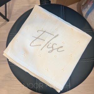 May include: Cream-colored baby blanket with the name "Elise" embroidered in a soft gray script. The blanket has a delicate, dotted pattern and a scalloped edge. The blanket is folded on a black leather stool.