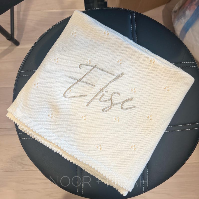 May include: Cream-colored baby blanket with the name "Elise" embroidered in a soft gray script. The blanket has a delicate, dotted pattern and a scalloped edge. The blanket is folded on a black leather stool.