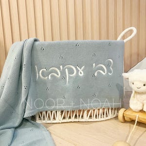 May include: A light blue baby blanket with embroidered Hebrew text is draped over a white woven basket. The basket has a handle and is filled with the blanket. A stuffed lamb toy on a wooden cart is in the background.
