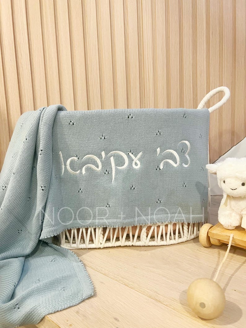 May include: A light blue baby blanket with embroidered Hebrew text is draped over a white woven basket. The basket has a handle and is filled with the blanket. A stuffed lamb toy on a wooden cart is in the background.