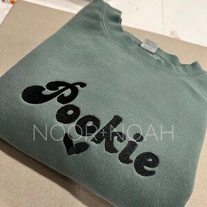 May include: A green sweatshirt with the word "cookie" embroidered in black on the front. The word is stylized with a rounded font.
