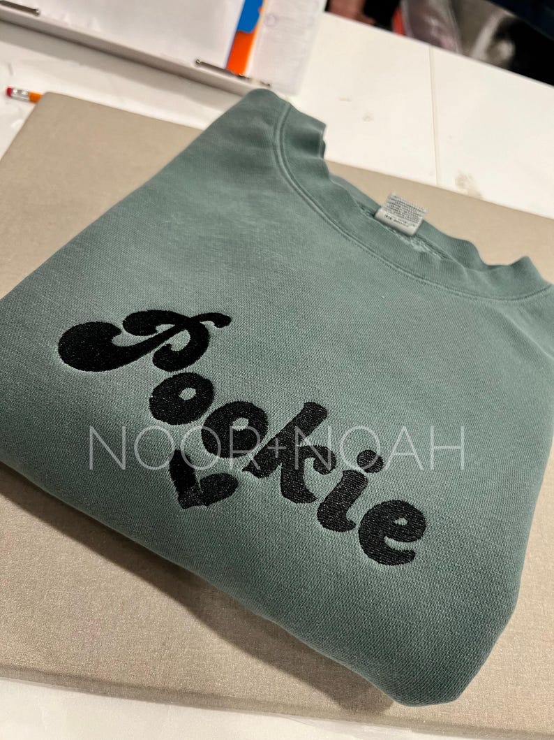 May include: A green sweatshirt with the word "cookie" embroidered in black on the front. The word is stylized with a rounded font.