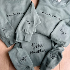 May include: Three green sweatshirts with black embroidered text. The top sweatshirt says "mama" with the words "ava" and "brennen" on the sleeves. The bottom sweatshirt says "twin mama" with the words "Lochlyn" and "Callan" on the sleeves. The middle sweatshirt says "mama" with the word "luco" on the sleeve.