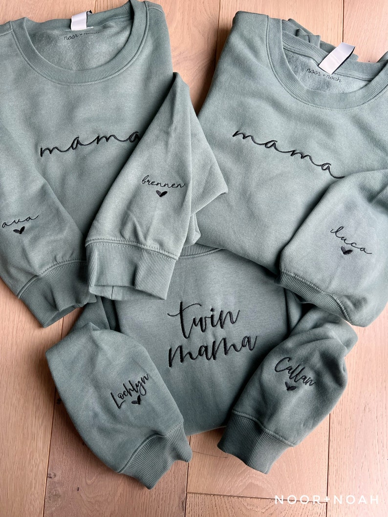 May include: Three green sweatshirts with black embroidered text. The top sweatshirt says "mama" with the words "ava" and "brennen" on the sleeves. The bottom sweatshirt says "twin mama" with the words "Lochlyn" and "Callan" on the sleeves. The middle sweatshirt says "mama" with the word "luco" on the sleeve.