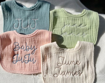 May include: Four personalized baby bibs in pastel colors: teal, light green, peach, and cream. Each bib features embroidered text with names or initials. The bibs are made of a soft, textured fabric, perfect for a baby shower gift.